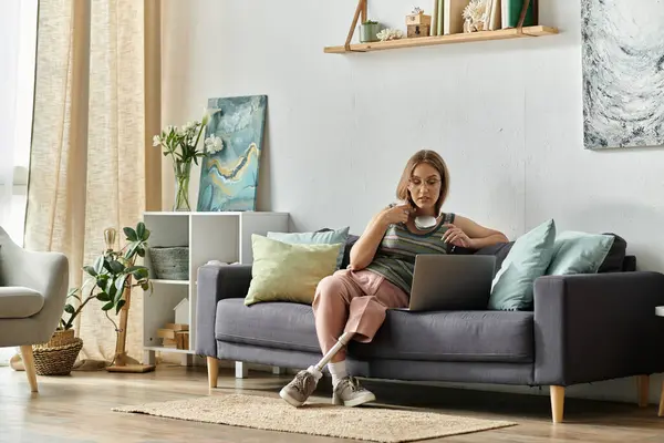 A young woman with a prosthetic leg sits on a couch in her modern living room, using a laptop. — Stock Photo