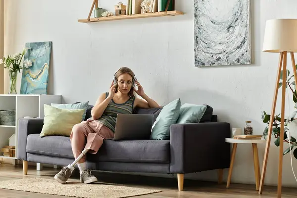 Una donna con una gamba protesica si siede su un divano con le cuffie, godendo la musica a casa — Foto stock