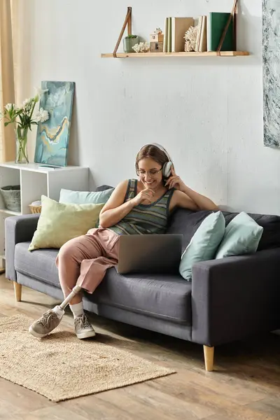 A young woman with a prosthetic leg sits on a couch in her living room, wearing headphones and smiling as she uses her laptop. — Stock Photo