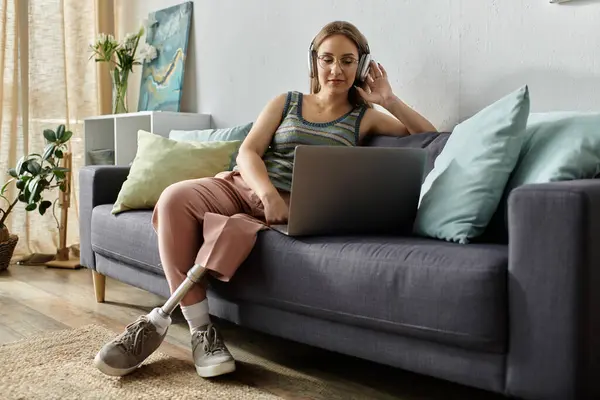 A young woman with a prosthetic leg relaxes on her living room couch, listening to music on her headphones and using a laptop. — Stock Photo
