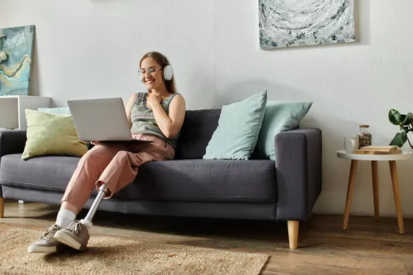 A young woman with a prosthetic leg smiles while using a laptop and wearing headphones on a gray couch in her modern living room. — Stock Photo