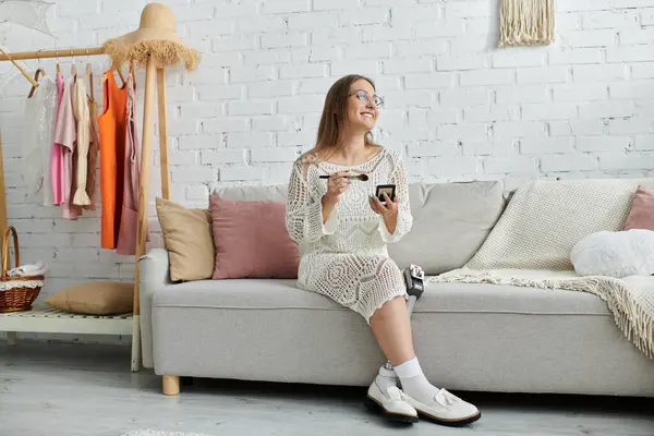 Una joven con una pierna protésica se sienta en un sofá, maquillándose con un cepillo y sonriendo — Stock Photo