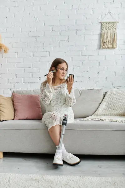 A young woman with a prosthetic leg sits on a couch, applying makeup while looking in a mirror. — Stock Photo