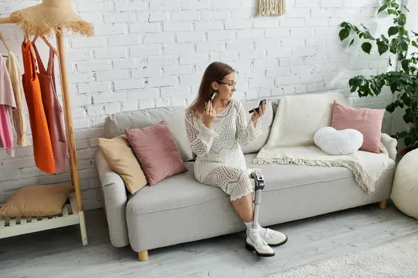 A young woman with a prosthetic leg sits on a couch and applies makeup, enjoying a moment of self-care at home. — Stock Photo
