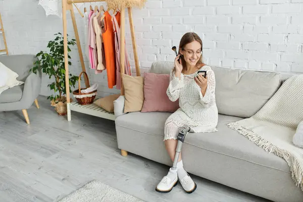 A young woman with a prosthetic leg sits on a couch in her home, applying makeup and smiling. — Stock Photo
