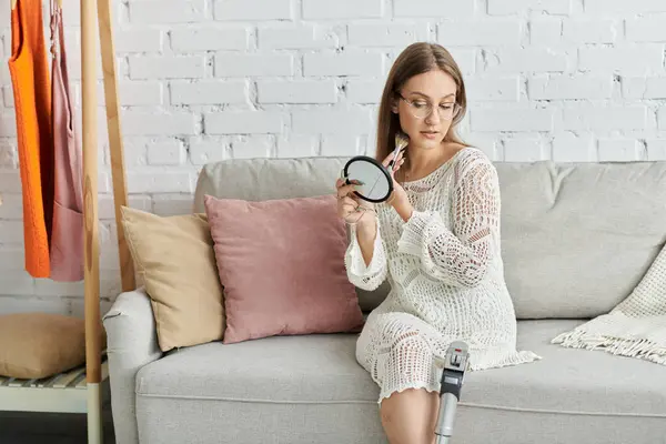 A young woman with a prosthetic leg sits on a couch, applying makeup with a brush and mirror. — Stock Photo