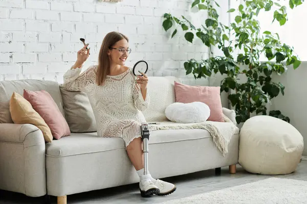 Uma jovem mulher com uma perna protética aplica maquiagem enquanto se senta em um sofá em sua sala de estar. — Fotografia de Stock