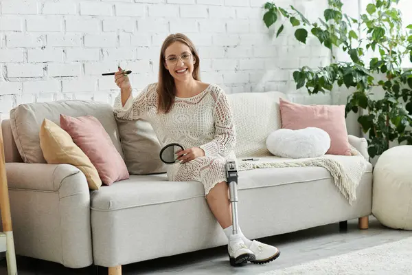 Une jeune femme avec une prothèse de jambe s'assoit sur un canapé, appliquant le maquillage tout en regardant un petit miroir. — Photo de stock