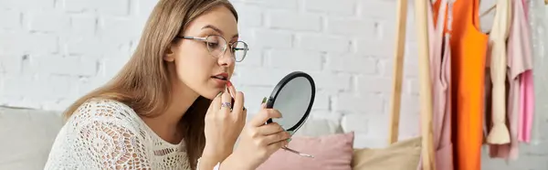 A young woman sits on a couch and applies lipstick while looking in a mirror. — Stock Photo