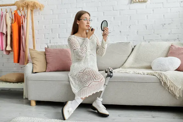 A young woman with a prosthetic leg sits on a couch, applying makeup while looking in a small mirror. — Stock Photo