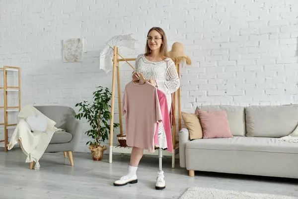 A young woman with a prosthetic leg stands in her living room, holding clothes from her closet. — Stock Photo