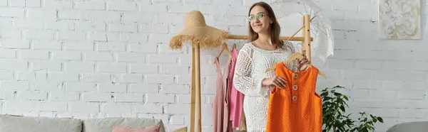 A young woman smiles as she holds up an orange dress in her home, surrounded by clothing and a plant. — Stock Photo