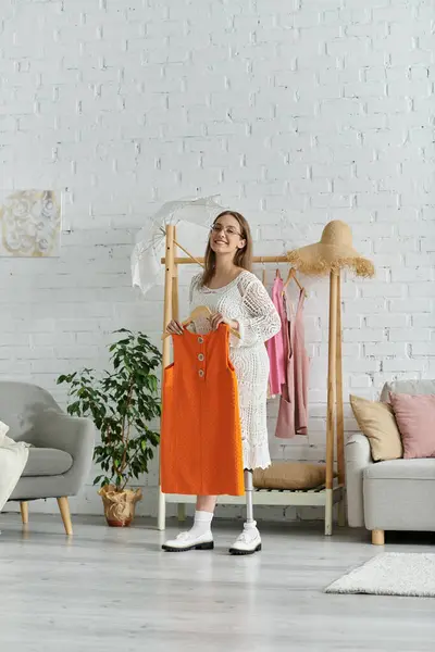 A young woman stands in her living room, smiling as she holds up an orange dress. — Stock Photo