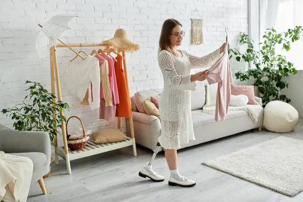 A young woman in a white dress stands in her living room, holding a pink garment and deciding what to wear for the day. — Stock Photo