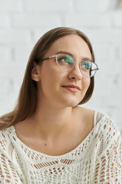 A young woman with long brown hair looks thoughtfully off-camera while wearing glasses and a white crocheted sweater. — Stock Photo