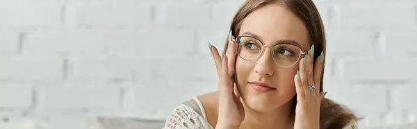 A young woman sits in her home, adjusting her glasses with a thoughtful expression. — Stock Photo