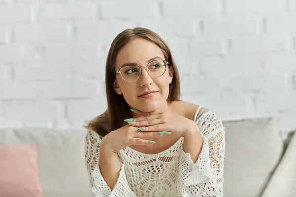 A young woman with glasses sits thoughtfully on a couch, lost in her own world. — Stock Photo