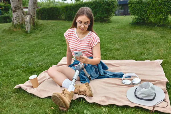 A young woman with a prosthetic leg sits on a blanket in a park, scrolling through her phone. — Stock Photo
