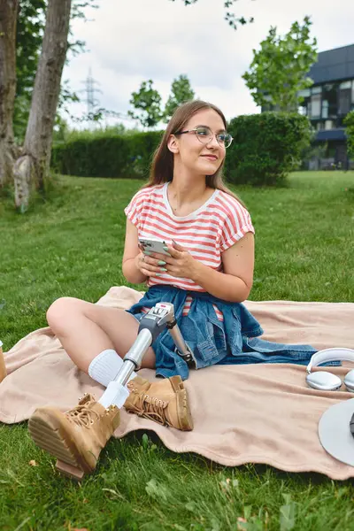 A young woman with a prosthetic leg relaxes on a blanket in a park, enjoying the sunshine and scrolling through her phone. — Stock Photo