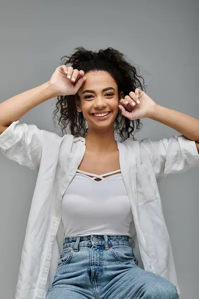 A young woman with curly hair smiles brightly as she poses in a white shirt against a grey background. — Stock Photo