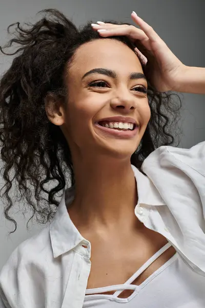 Una joven afroamericana con el pelo rizado, vistiendo una camisa blanca, sonríe brillantemente sobre un fondo gris. - foto de stock