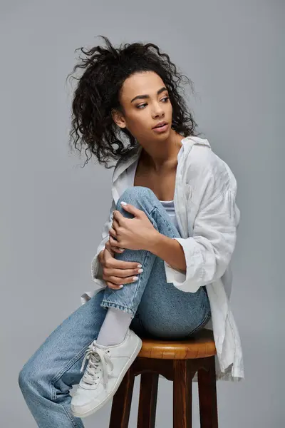 A young woman with curly hair sits on a stool against a gray backdrop. — Stock Photo