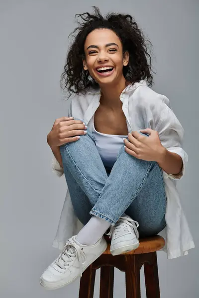 Una bella giovane donna nera con i capelli ricci siede su uno sgabello, sorridendo brillantemente alla macchina fotografica. — Foto stock