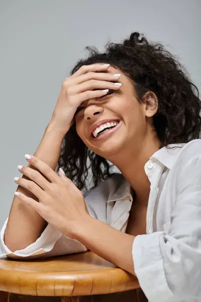 Une jeune femme aux cheveux bouclés rit chaleureusement, couvrant son visage de sa main, assise sur un tabouret en bois sur fond gris. — Photo de stock