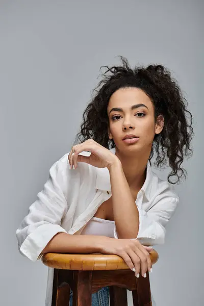 A young woman with curly hair, wearing a white shirt, rests her arm on a wooden stool against a grey backdrop. — Stock Photo