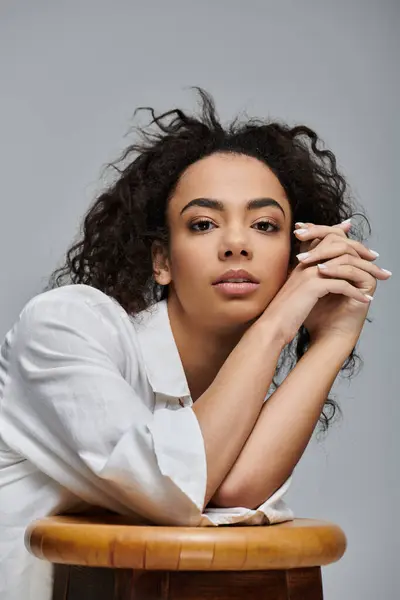 A beautiful young woman with dark curly hair leans forward, resting her arms on a wooden stool against a grey background. — Stock Photo
