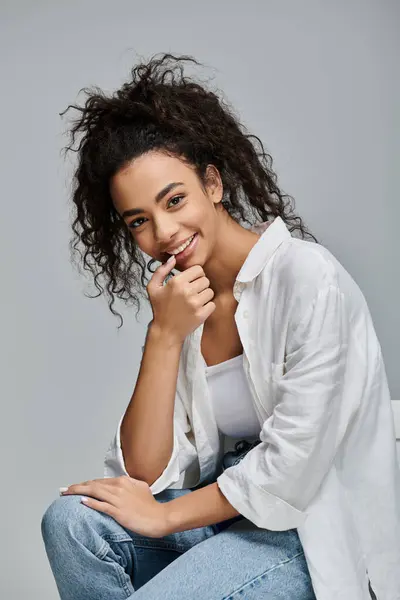 Une jeune femme aux cheveux bouclés sourit en toute confiance, portant une chemise blanche et un jean, sur un fond gris. — Photo de stock