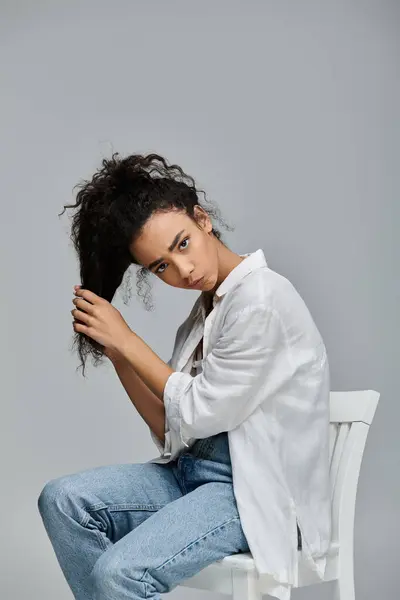 A young Black woman with curly hair sits on a white chair against a gray background, looking confident and beautiful. — Stock Photo