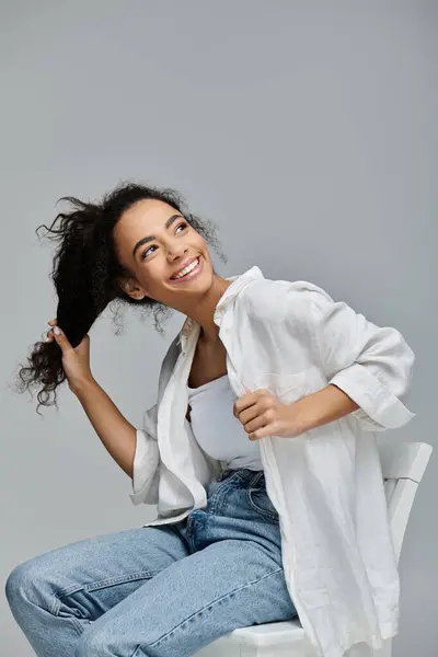 A young Black woman sits on a white chair against a gray backdrop, playfully tossing her hair while beaming with joy. — Stock Photo