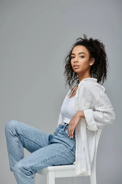 A young African American woman poses confidently in a white shirt and blue jeans, sitting on a chair against a grey background. — Stock Photo
