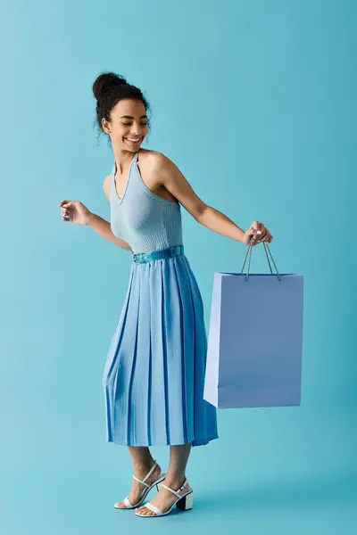 A smiling young woman in a blue dress holds a shopping bag while walking on a blue background. — Stock Photo