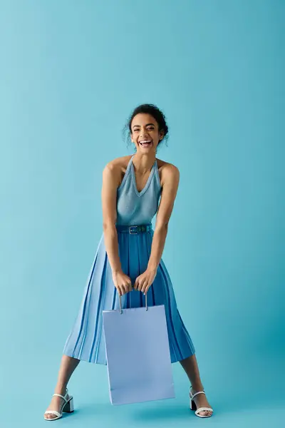 Uma bela jovem mulher em um vestido azul, sorri com um grande saco de compras azul na frente de um fundo azul brilhante. — Fotografia de Stock
