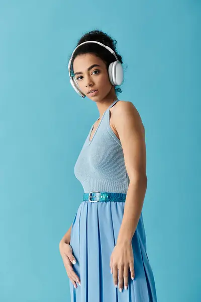 Una joven con un vestido azul se levanta contra un fondo azul brillante, auriculares puestos, perdidos en la música. - foto de stock