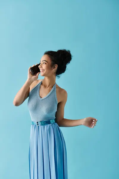 Una giovane donna afroamericana con un vestito blu sorride mentre parla al telefono su uno sfondo blu vibrante. — Foto stock