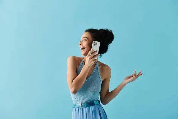 A young African American woman in a blue dress smiles brightly while talking on her phone against a light blue background. — Stock Photo