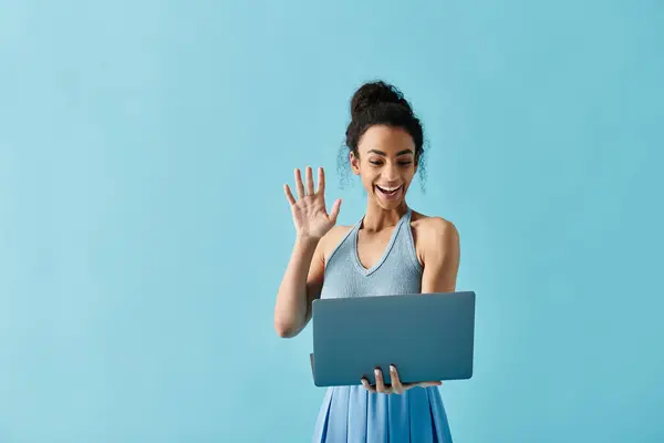 A young woman in a blue dress waves with a laptop in hand, standing against a light blue backdrop. — Stock Photo