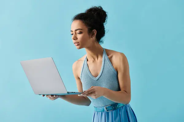 Una joven afroamericana con un vestido azul sostiene un portátil y mira hacia abajo a la pantalla. — Stock Photo