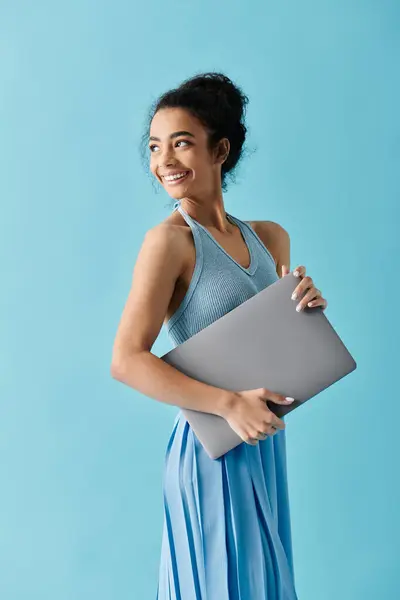 A young African American woman in a blue dress smiles confidently while holding a laptop against a blue background. — Stock Photo