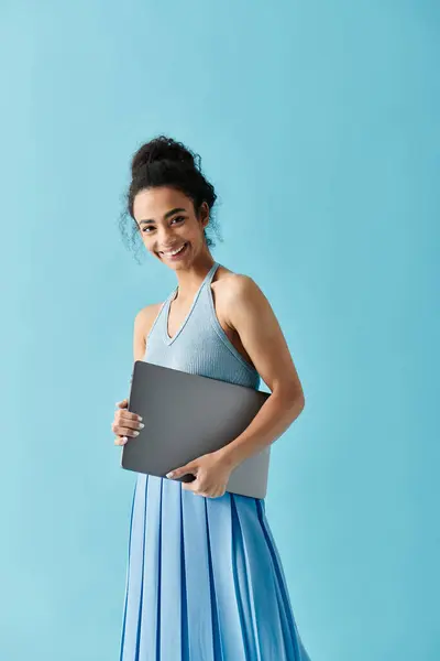 A young African American woman smiles as she holds a laptop in front of a blue background. — Stock Photo