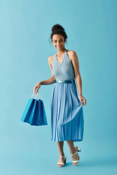 A young woman in a blue dress smiles brightly as she carries blue shopping bags against a vibrant blue background. — Stock Photo