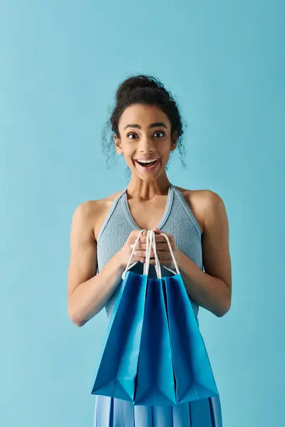 Uma jovem mulher negra segura sacos de compras azuis contra um fundo azul claro, seu rosto mostrando alegria — Fotografia de Stock