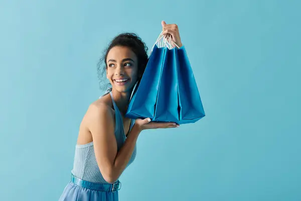 Uma jovem mulher em um vestido azul sorri brilhantemente enquanto segura uma pilha de sacos de compras azuis. — Fotografia de Stock