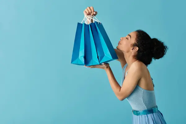 Uma jovem mulher em um vestido azul excitadamente segura uma pilha de sacos de compras azuis contra um fundo azul claro. — Fotografia de Stock