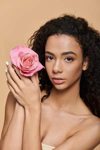 A young African American woman with natural makeup holds a pink rose while looking at the camera. — Stock Photo