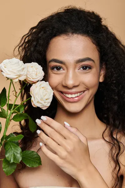 Una giovane donna afroamericana con le spalle nude e il trucco naturale sorride tenendo in mano rose bianche. — Foto stock