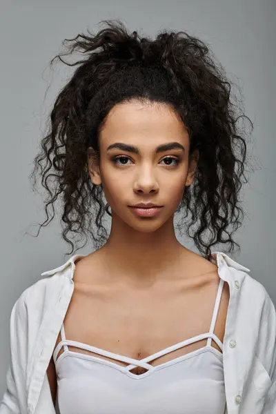 A young woman with dark curly hair looks confidently at the camera, wearing a white top and a white shirt unbuttoned over it. — Stock Photo